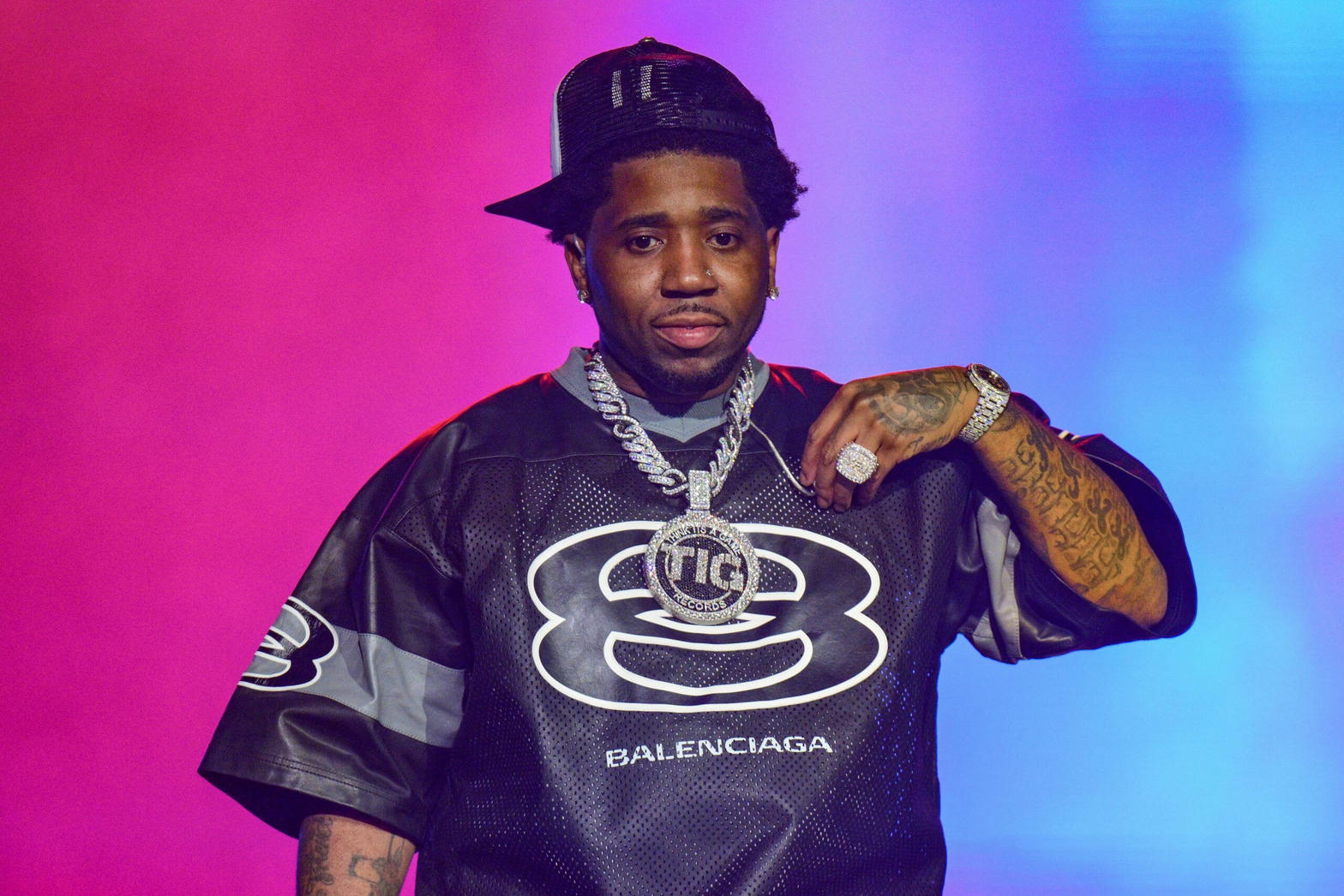 YFN Lucci on stage in a black jersey and Think It's a Game chain, pink and blue light behind him
